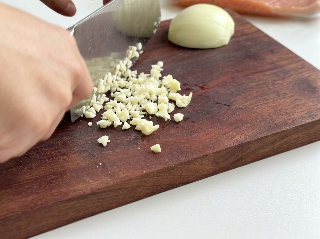 A close-up shot of a person using a chefs knife to finely chop white onion and garlic on a wooden board.