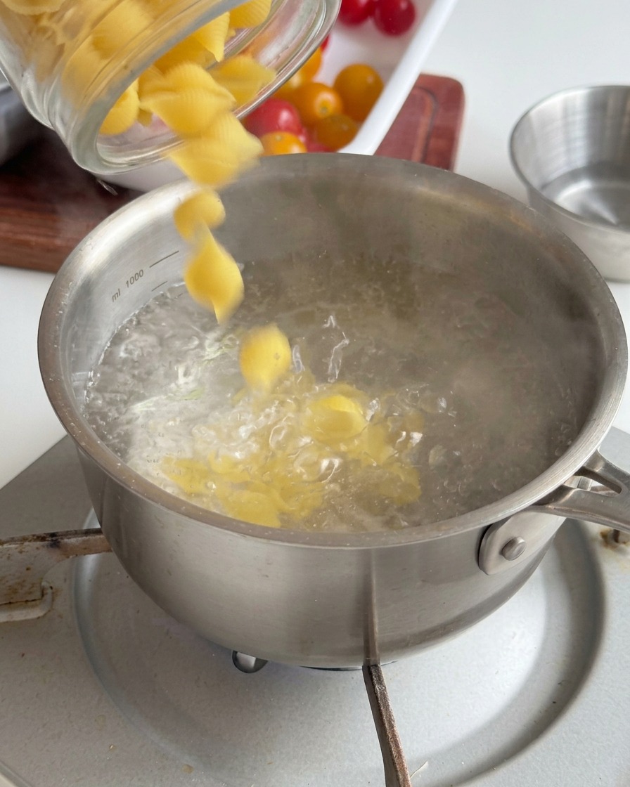 Yellow shell-shaped pasta being poured from a glass jar into a stainless steel pot of bubbling boiling water.