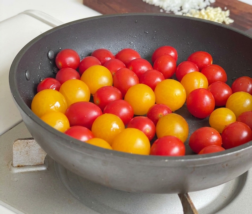 A mix of red and yellow cherry tomatoes being sautéed in a dark frying pan on a gas stove.
