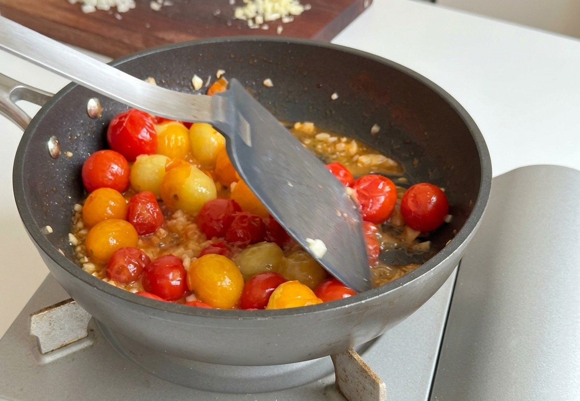 Minced garlic and onion being stirred into a pan of softened red and yellow cherry tomatoes with a metal spatula.