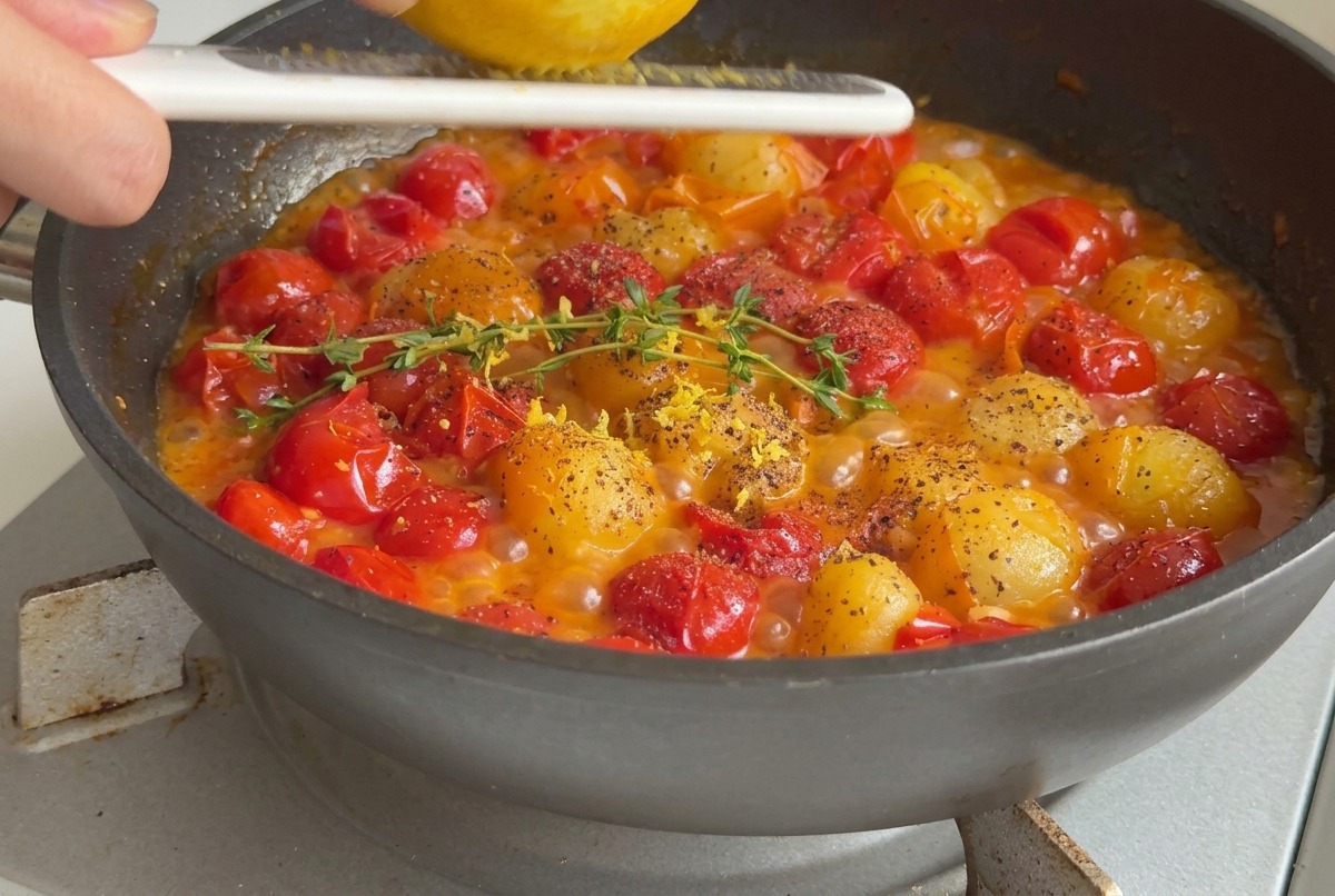 A hand grating lemon zest over a pan of bubbling cherry tomatoes seasoned with fresh thyme and black pepper.