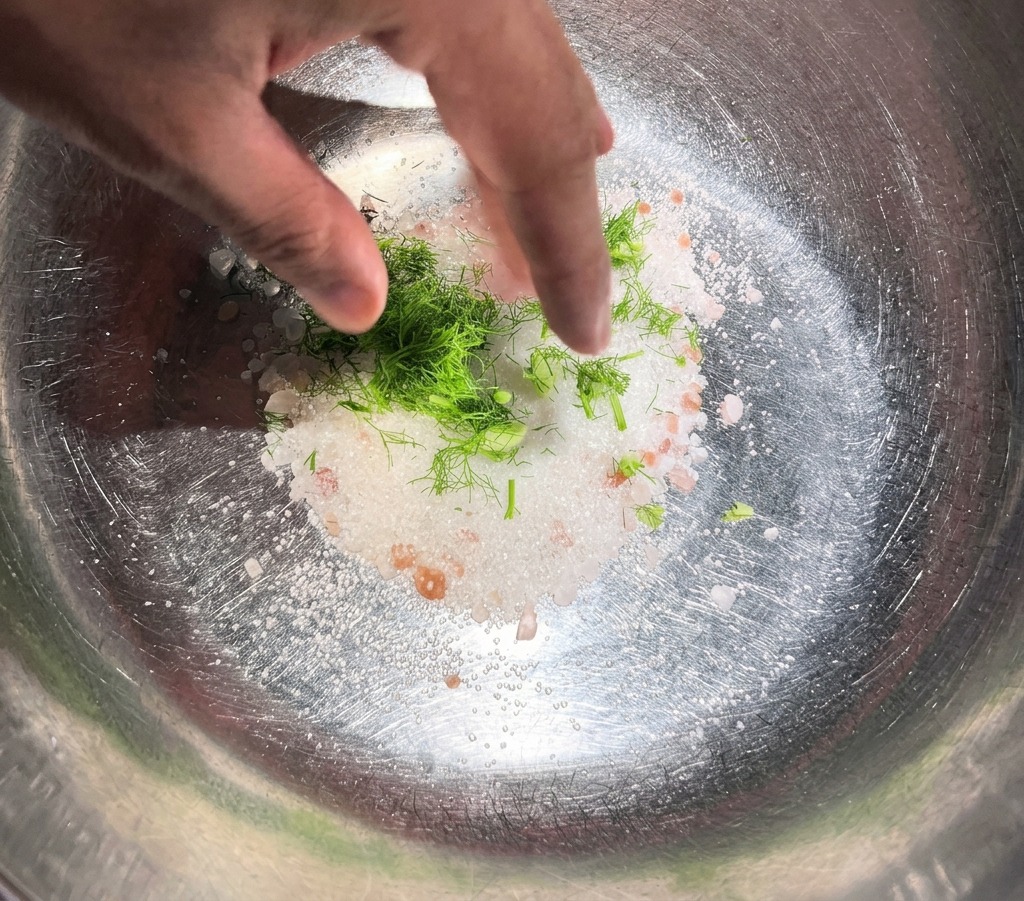 A hand mixing fresh green dill and whole spices into a pile of white salt and sugar inside a stainless steel bowl.