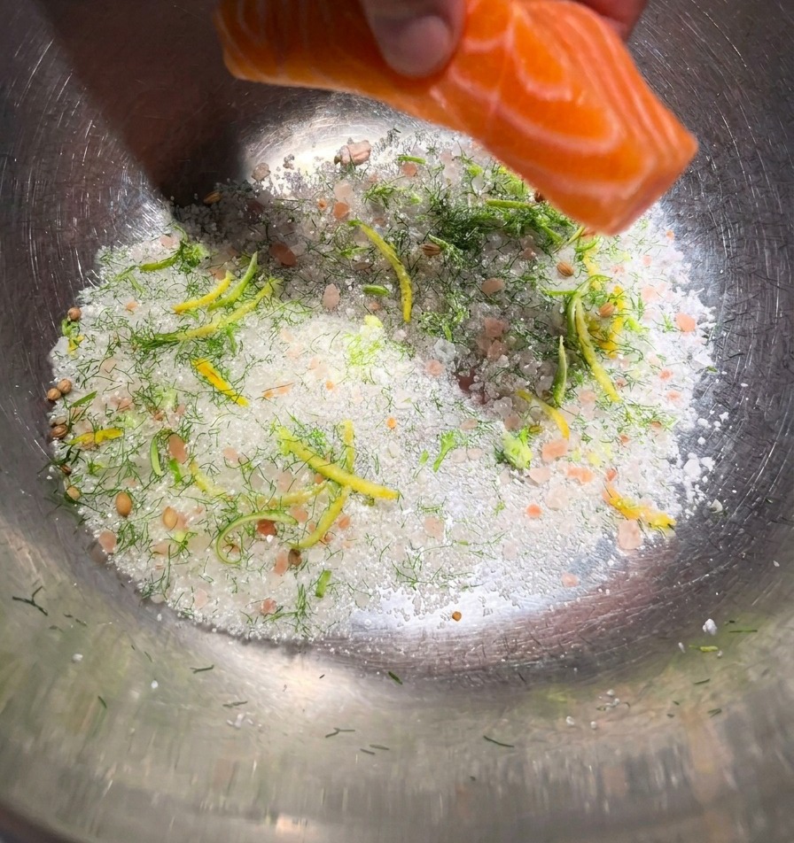 A hand lowering a vibrant orange salmon fillet onto a bed of curing salts and green herbs in a metal mixing bowl.