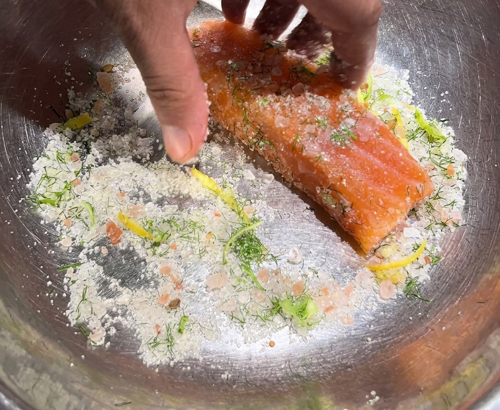 Hands rubbing a coarse mixture of salt, sugar, and herbs onto a thick piece of raw salmon in a metal bowl.