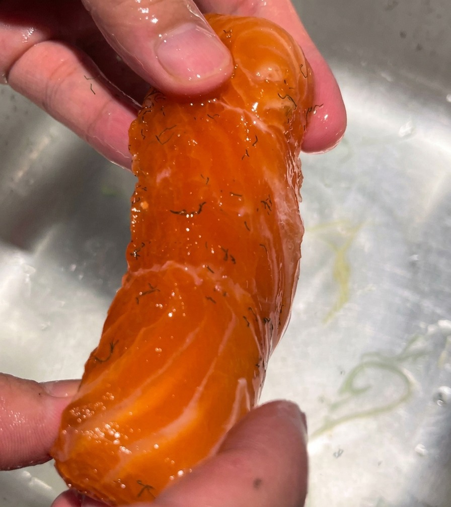 A hand holding a cured salmon fillet under a stream of running water to wash off the salt and spice mixture.