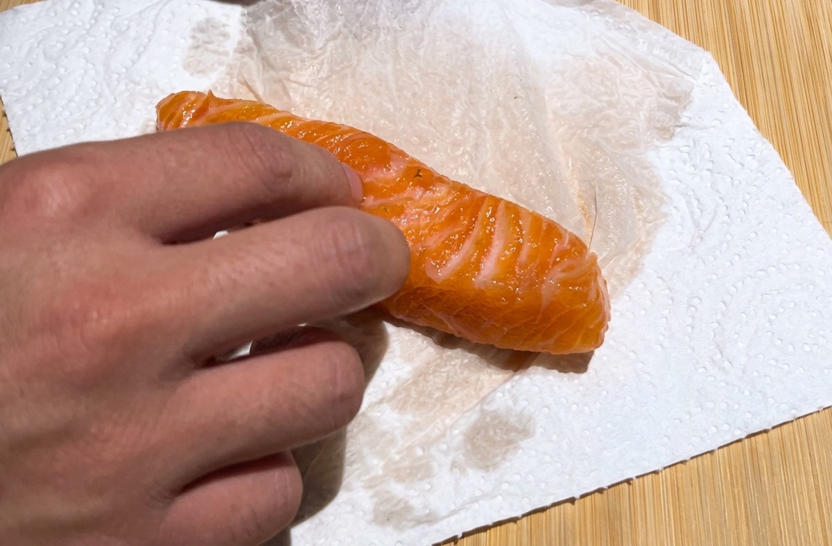 A hand using a white paper towel to pat dry a bright orange cured salmon fillet on a wooden board.