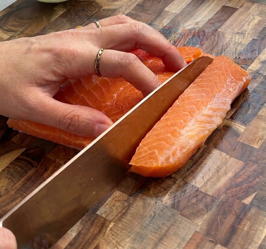 A close-up shot of hands using a knife to cut a large slab of raw salmon into small cubes on a wooden board.