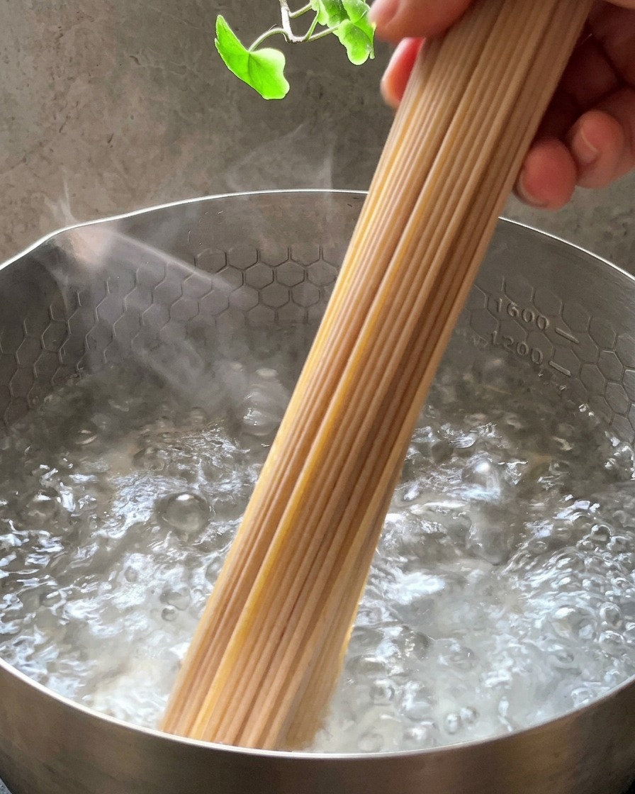 A bundle of dry whole wheat spaghetti being dipped into a stainless steel pot of bubbling boiling water.