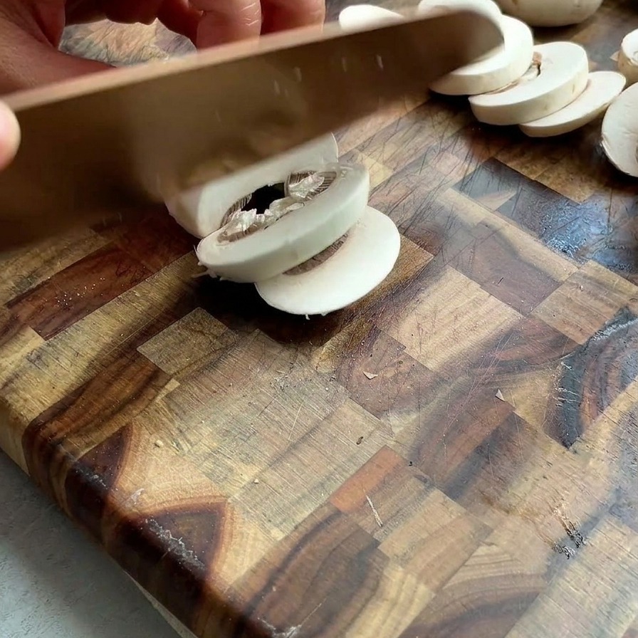 Hands slicing fresh white mushrooms into thin slices on a wooden butcher-block cutting board.