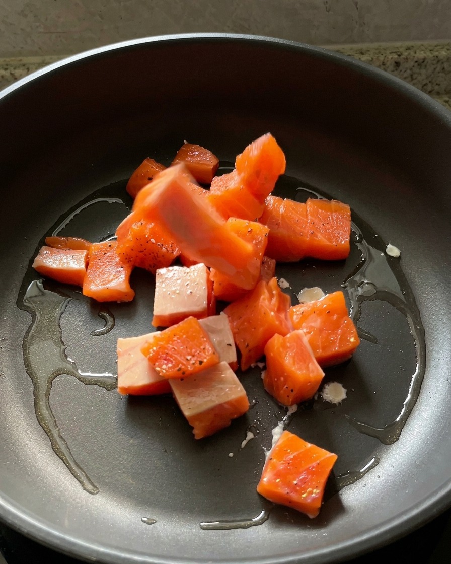 Marinated raw salmon cubes being placed into a dark skillet with shimmering hot oil.
