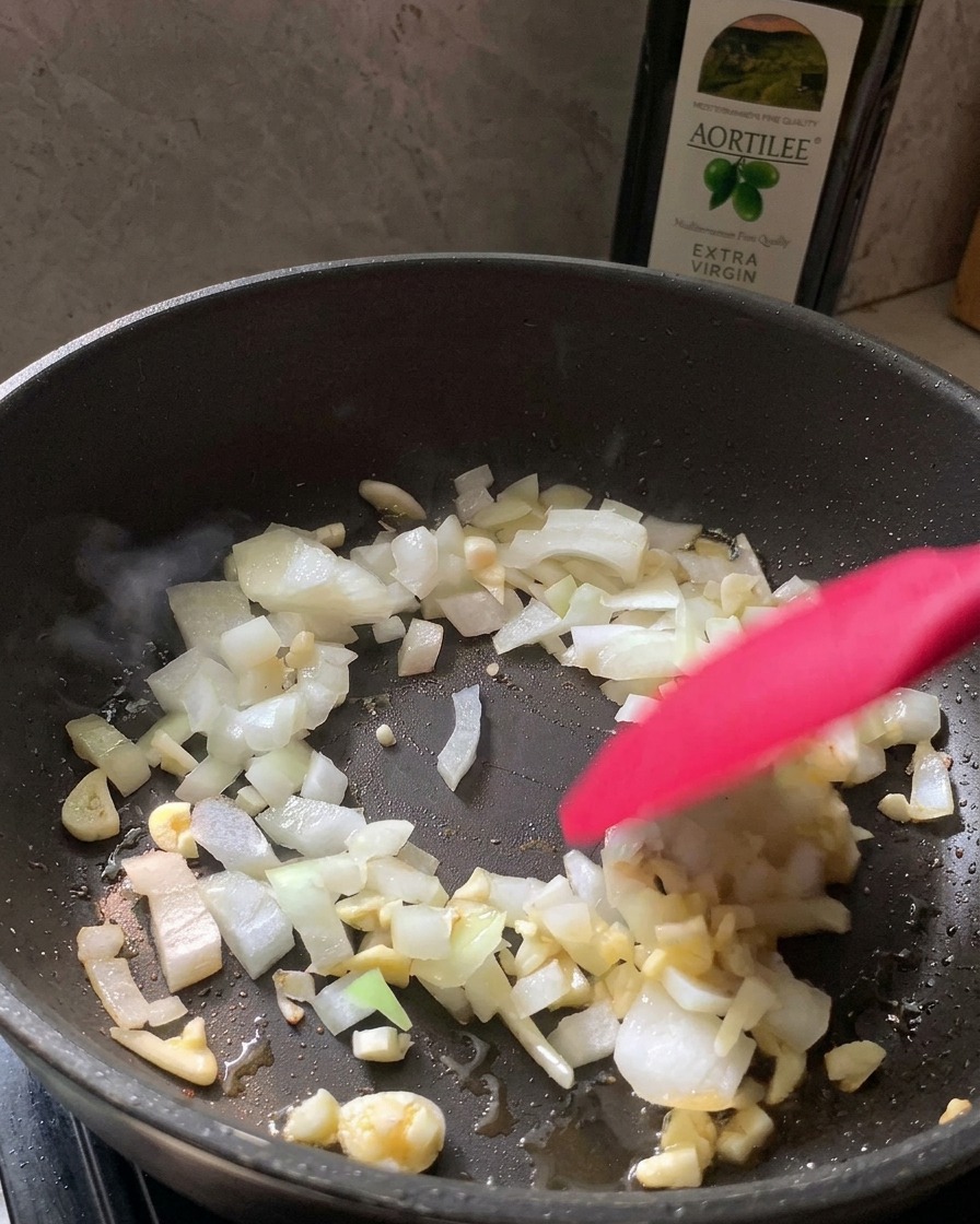 A red spatula stirring chopped white onions and minced garlic in a black frying pan.