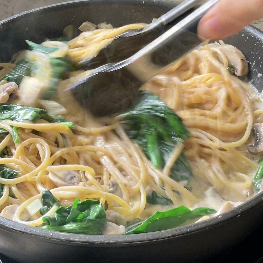 Tongs tossing brown spaghetti noodles into a white cream sauce with green spinach leaves in a black frying pan.