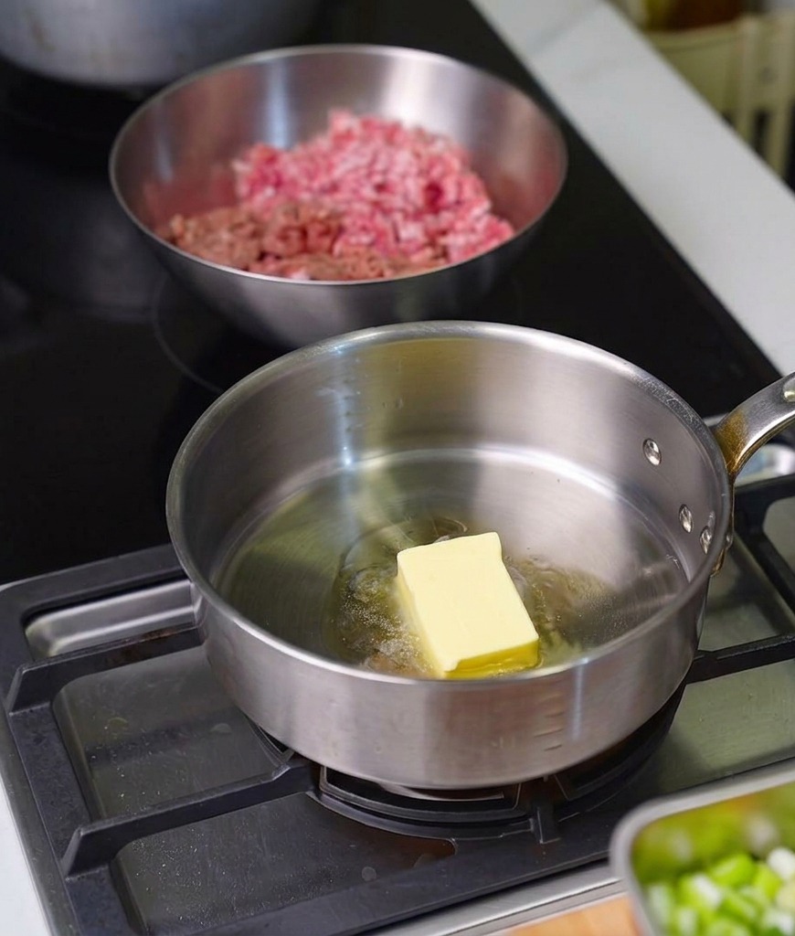 A block of yellow butter melting into a pool of oil in a shiny stainless steel saucepan on a stovetop.