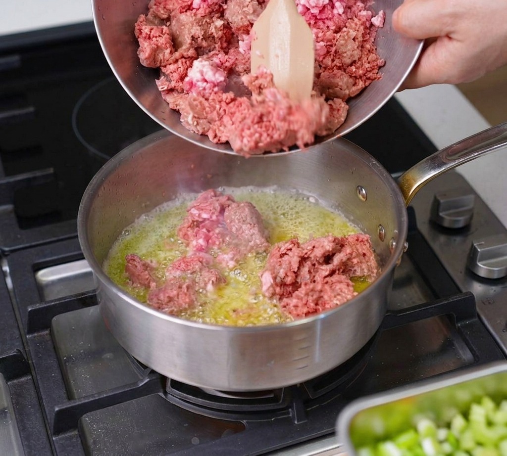 Ground beef and pork being poured from a metal bowl into a sizzling pan with melted butter.