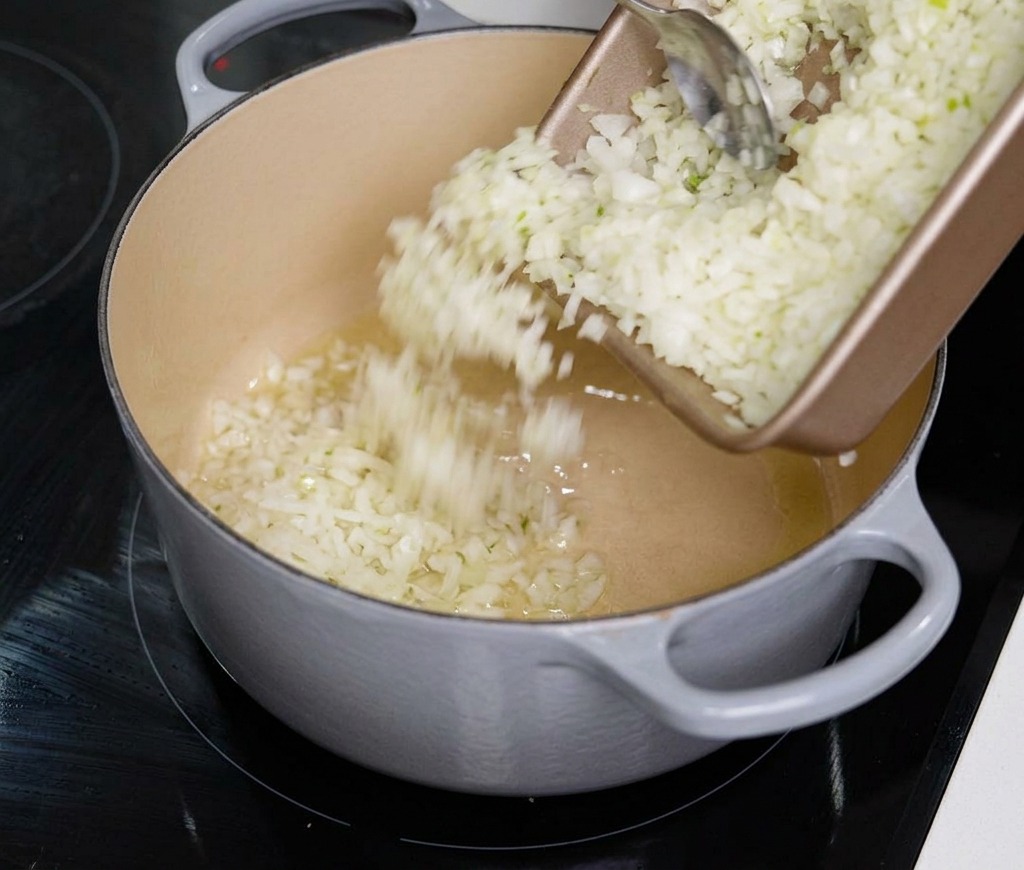 Finely chopped white onions being poured from a rectangular tray into a large grey Dutch oven.