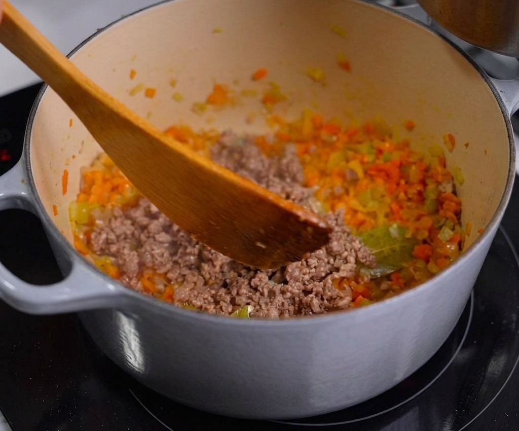 A person using a wooden spatula to stir browned minced meat into a pot of sautéed diced vegetables.
