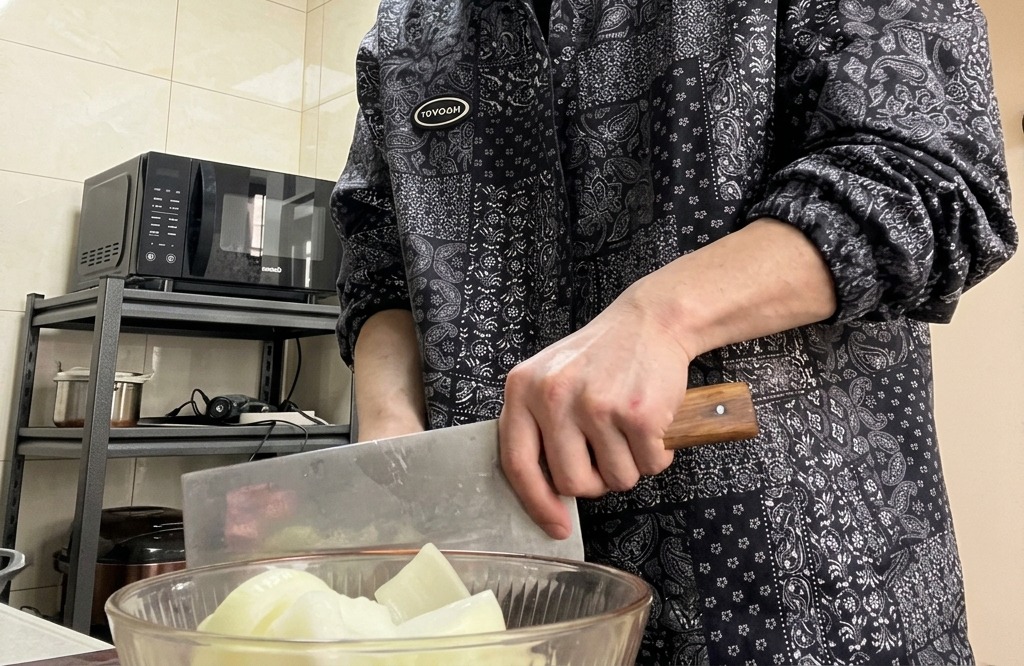 Hands holding a large cleaver-style knife, chopping an onion on a wooden cutting board.