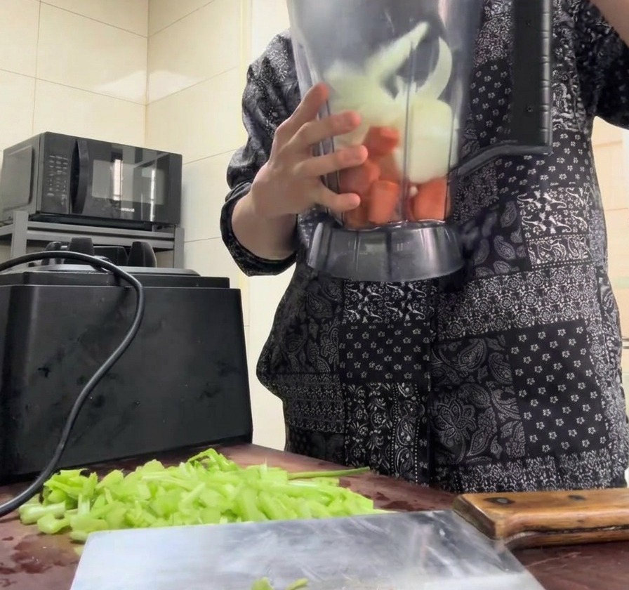 Chopped carrots, onions, and celery being poured into a clear blender pitcher.