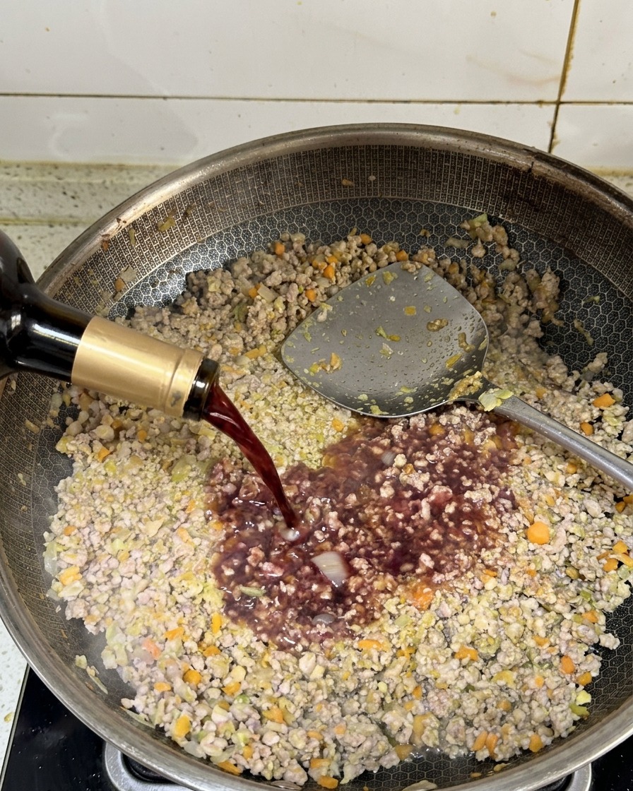 Dark red wine being poured from a bottle into a pan of browned ground meat.