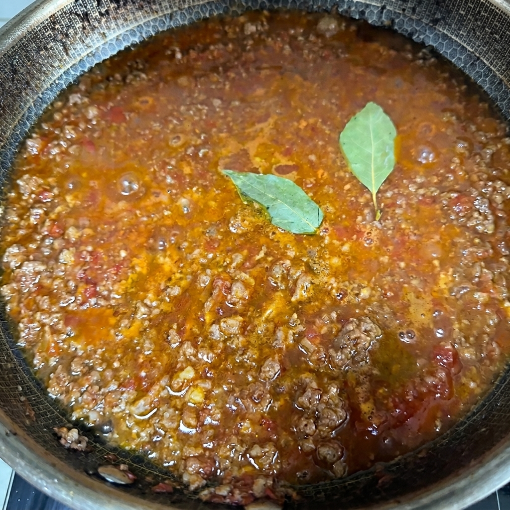 Close-up of a bubbling, rich meat sauce simmering in a large pan with two whole green bay leaves resting on the surface.
