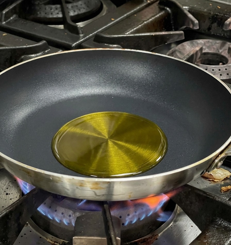 Olive oil shimmering in the center of a black non-stick frying pan over a blue gas flame.