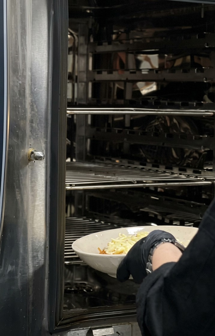 A chef in black gloves sliding a white bowl of cheese-topped pasta into a large industrial stainless steel oven.