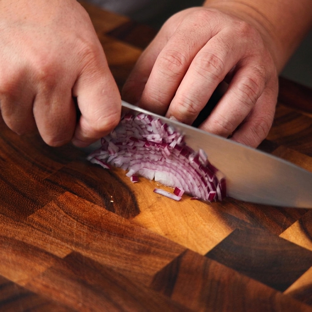 Close-up of hands using a chefs knife to finely dice a purple onion on a wooden cutting board.