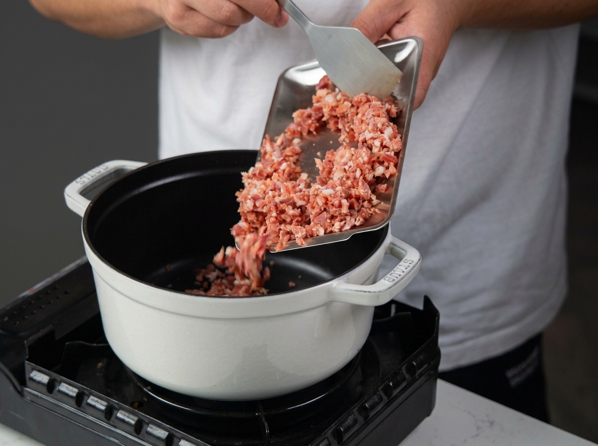 Pouring diced pancetta from a metal tray into a white enameled cast-iron pot on a portable stove.
