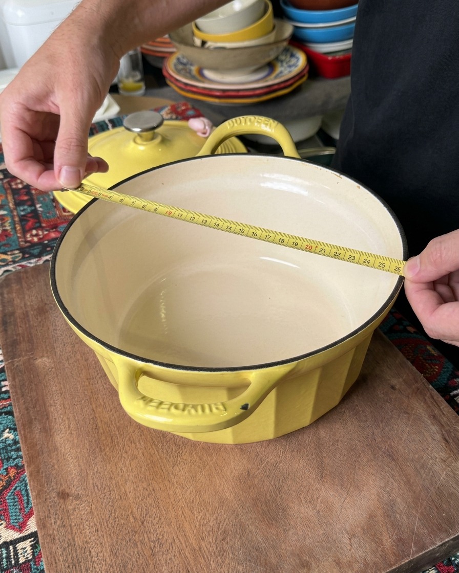 A person measuring the top diameter of a yellow Dutch oven with a yellow measuring tape to confirm it is roughly 26 centimeters wide.