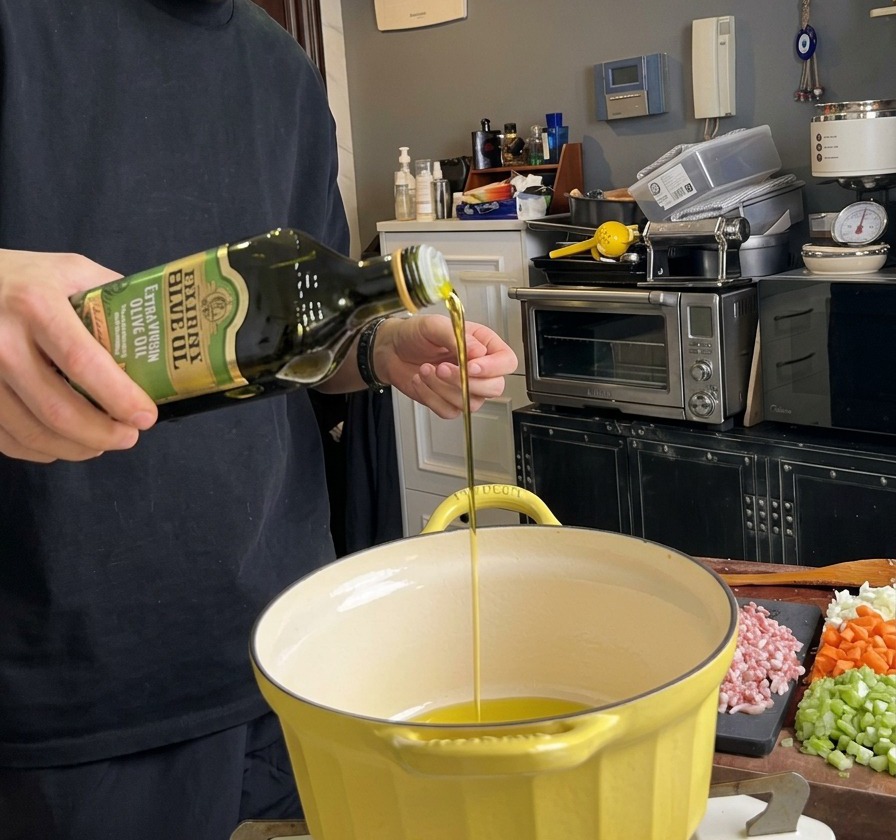 Olive oil being poured from a green bottle into a heated yellow pot, with chopped bacon resting on a cutting board nearby.