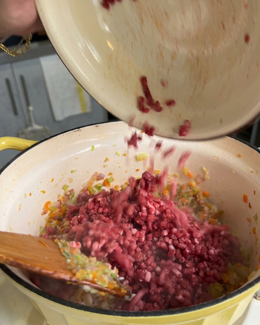Ground meat being poured from a bowl into a pot filled with sautéed diced carrots, celery, and onions.