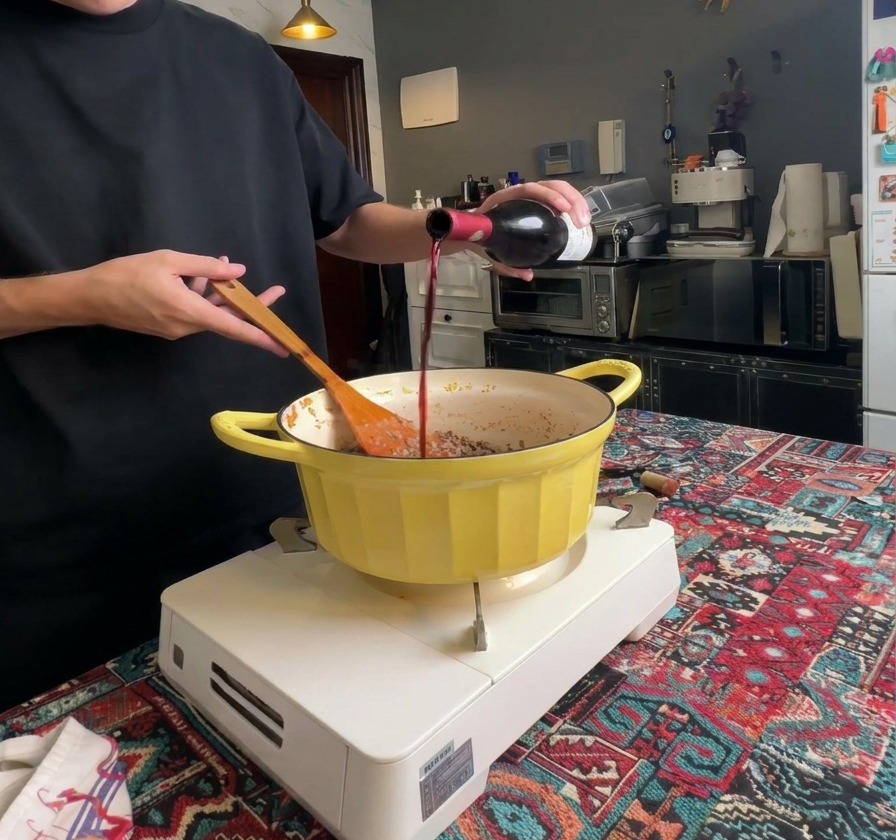 Red wine being poured from a dark glass bottle into a pot filled with well-browned ground meat.