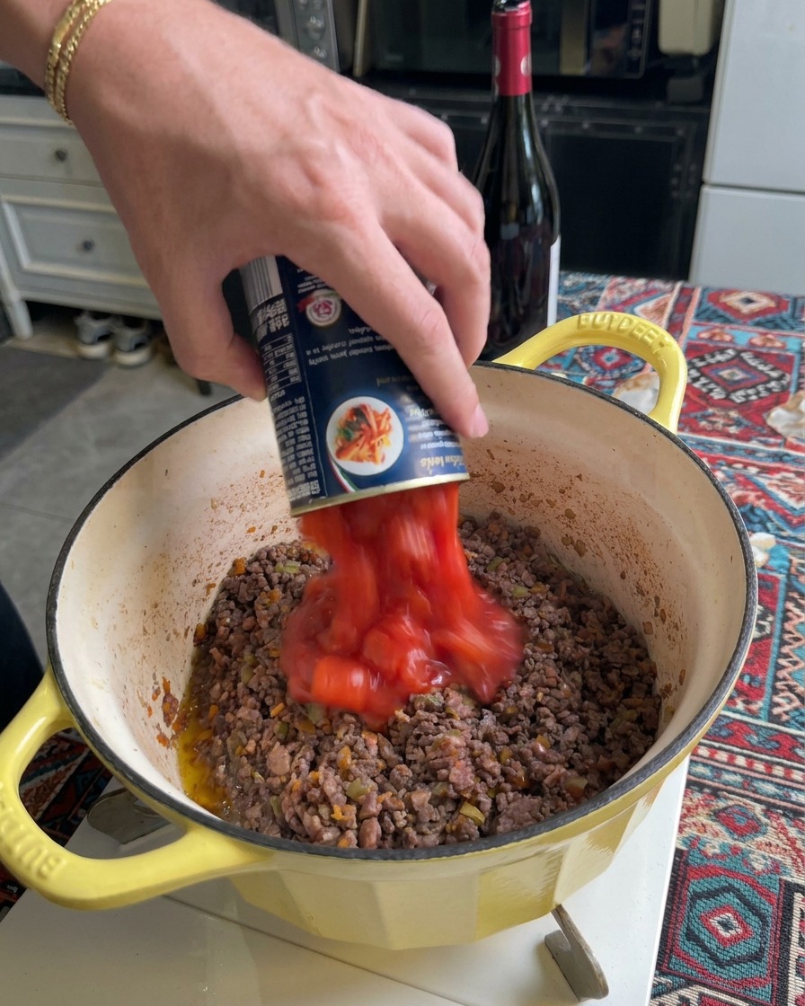 Canned diced tomatoes and puree being poured into a pot of browned meat.