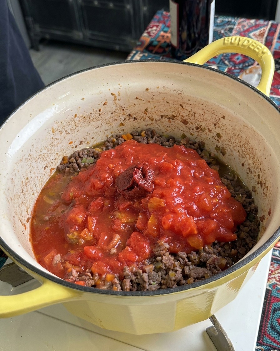 A dollop of thick, dark tomato paste sitting on top of crushed tomatoes and ground meat in a yellow pot.