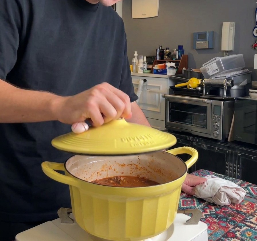 A person placing a yellow cast iron lid onto a pot of ragù to begin the slow simmering process.