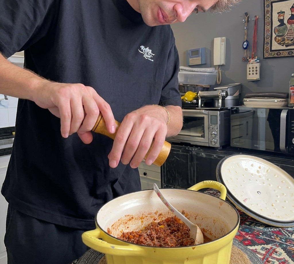 A person using a wooden pepper grinder to add fresh black pepper to a thick Italian meat sauce in a yellow Dutch oven.