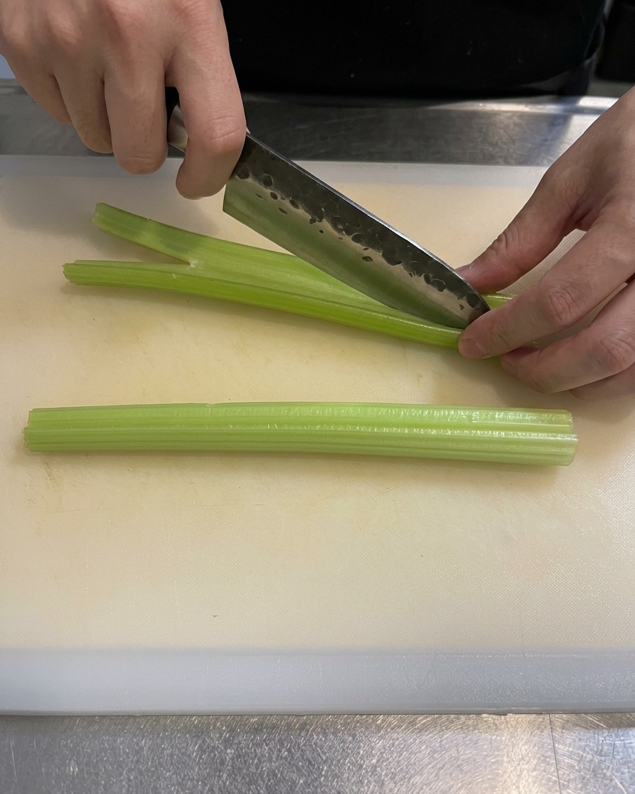 A close-up of a chefs hand using a sharp knife to slice green celery stalks into small pieces on a clean white cutting board.