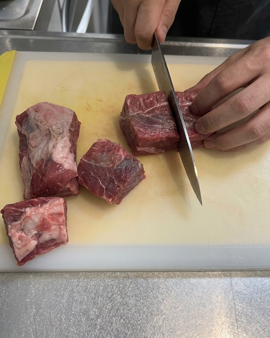 Raw marbled beef brisket being cut into large square chunks on a white cutting board.