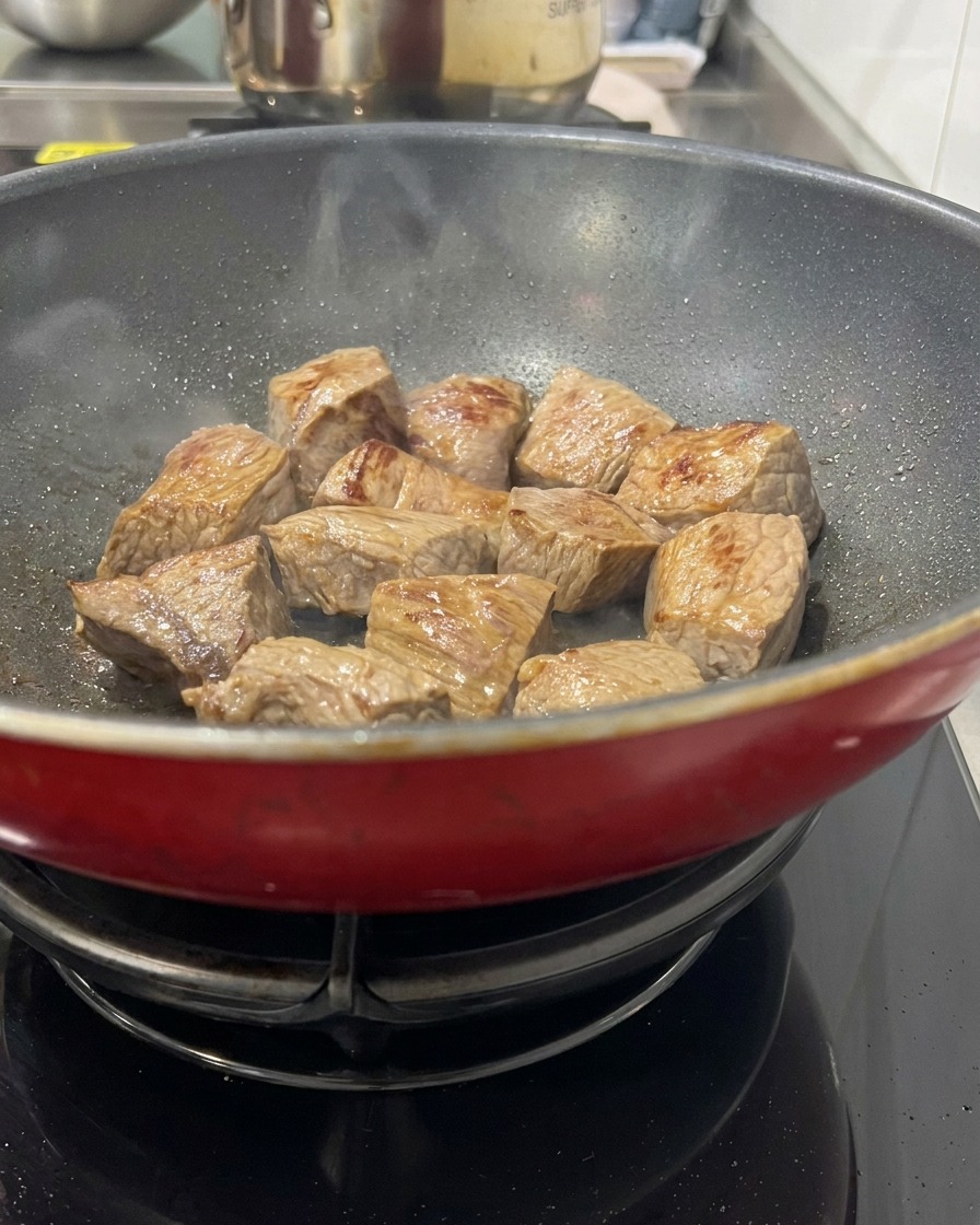 Cubed beef browning in a hot red pan, showing seared golden surfaces and light steam rising from the meat.