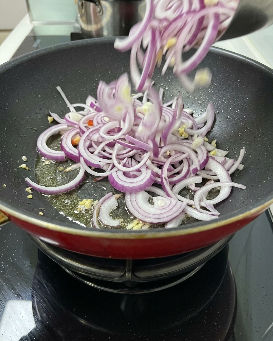 A large quantity of sliced red onions being poured from a bowl into a frying pan with oil and aromatics to begin sautéing.