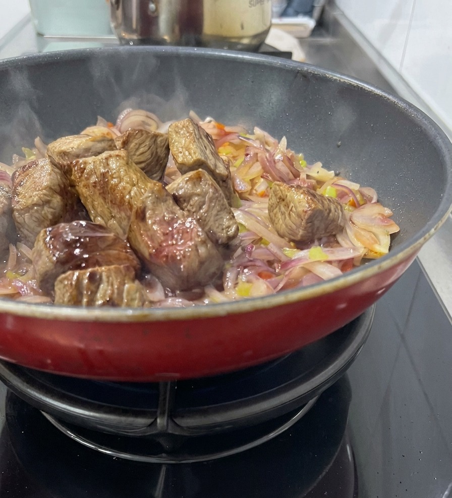 Seared beef cubes sitting on a bed of sautéed red onions and celery in a red pan, with wine being added to deglaze.