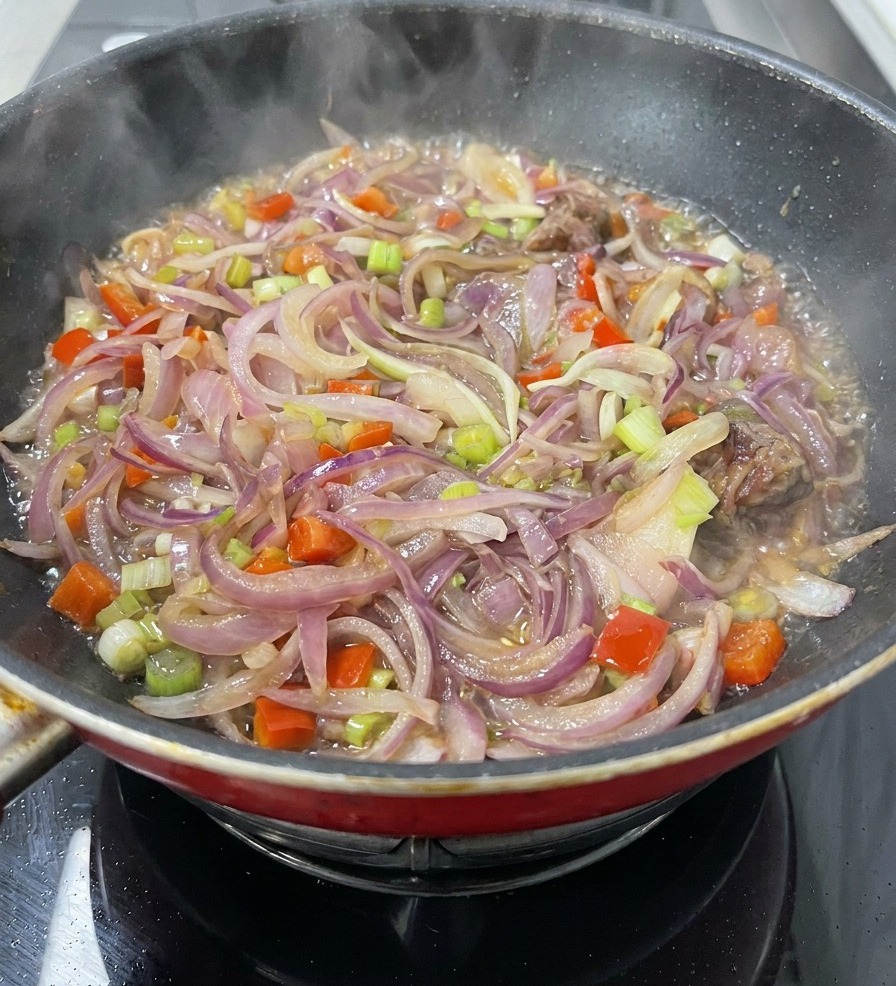 A pan filled with sliced red onions, carrots, celery, and meat pieces simmering in a light liquid with steam rising.