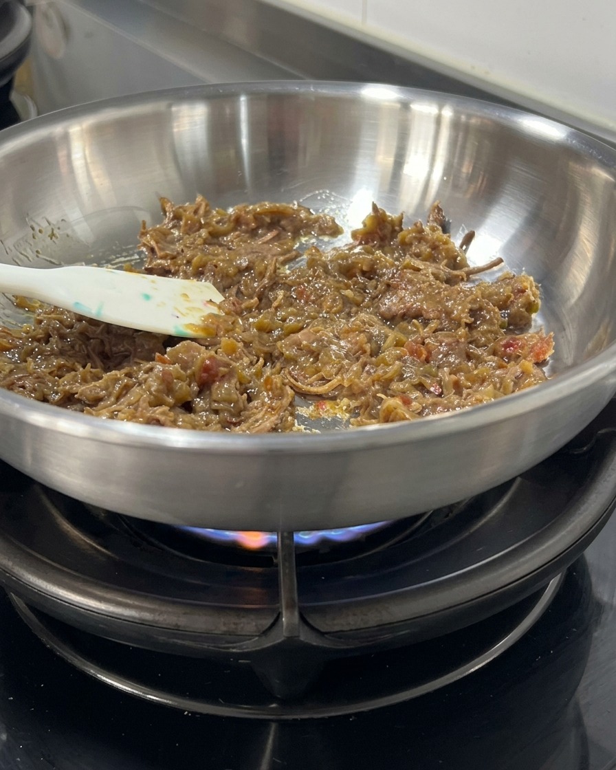 Thick, dark meat sauce and shredded beef being stirred with a spatula in a stainless steel skillet over a flame.