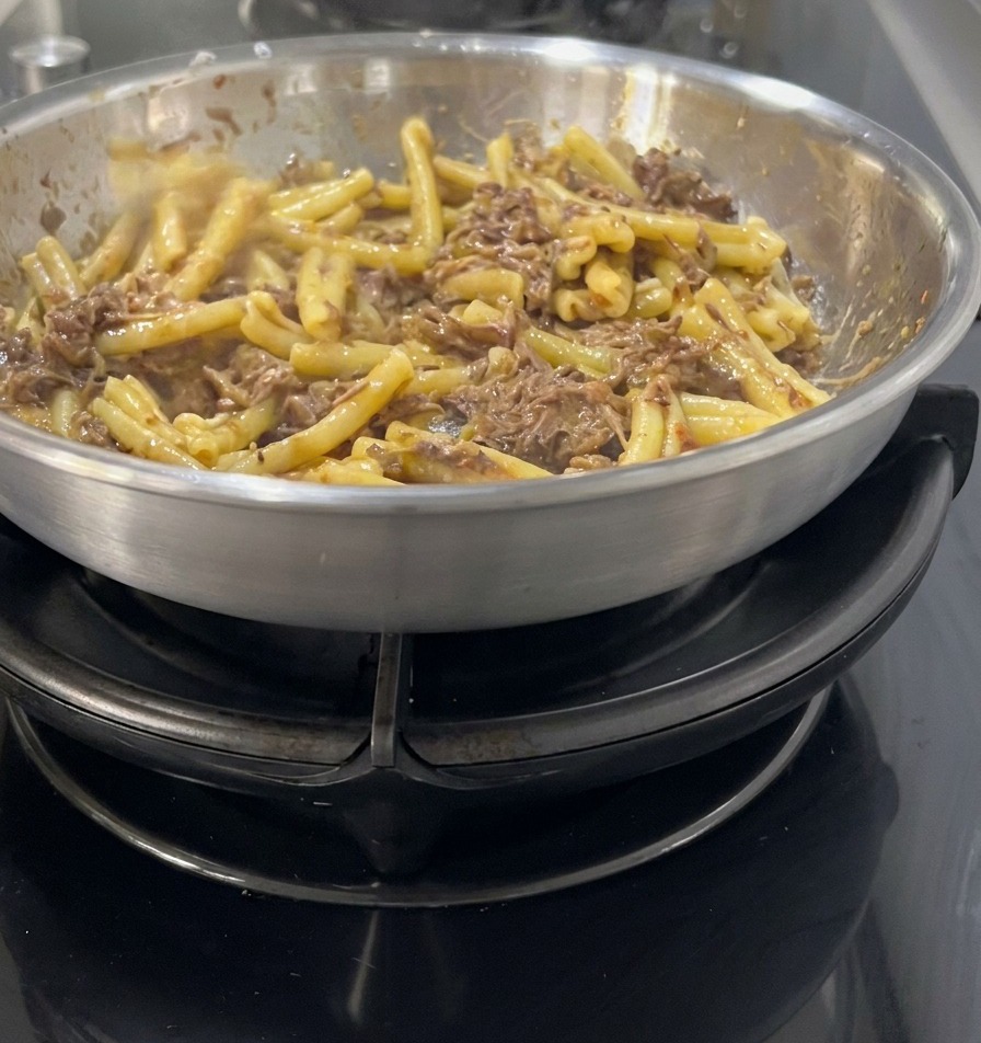 Casarecce pasta being folded into a dark, rich shredded meat and onion sauce in a stainless steel skillet.