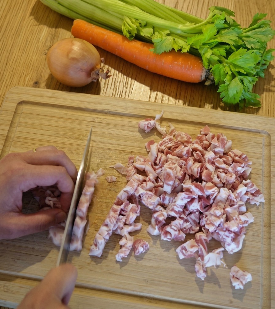 A close-up shot of hands using a chefs knife to dice pink and white strips of pancetta into small cubes on a light-colored wooden cutting board.