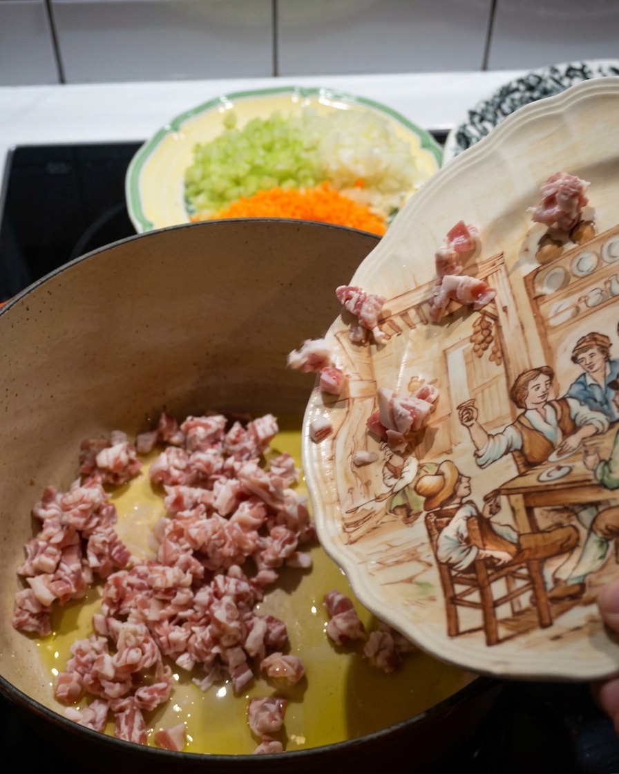 Diced pancetta cubes being poured from a decorative ceramic plate into a hot red cast-iron pot containing olive oil.