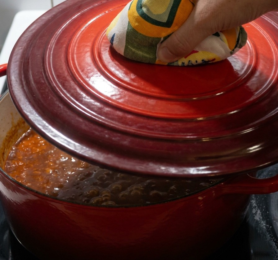 A hand wearing a yellow and green patterned oven mitt placing a red cast-iron lid onto a simmering pot.