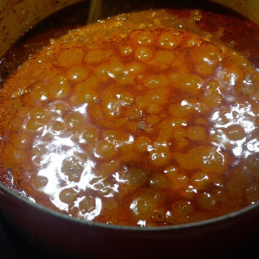 Ground nutmeg being sprinkled over the top of a thick, orange-hued meat sauce simmering in a pot.