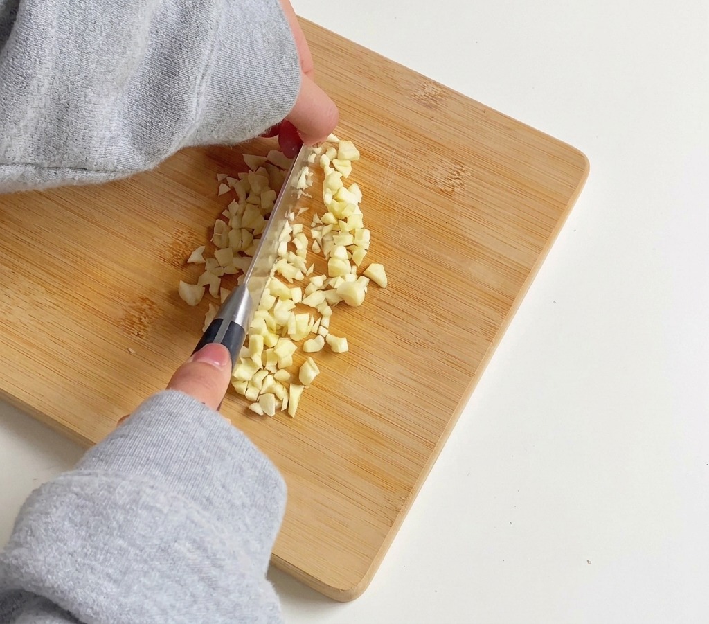A person using a silver kitchen knife to finely mince several cloves of garlic on a bamboo cutting board.
