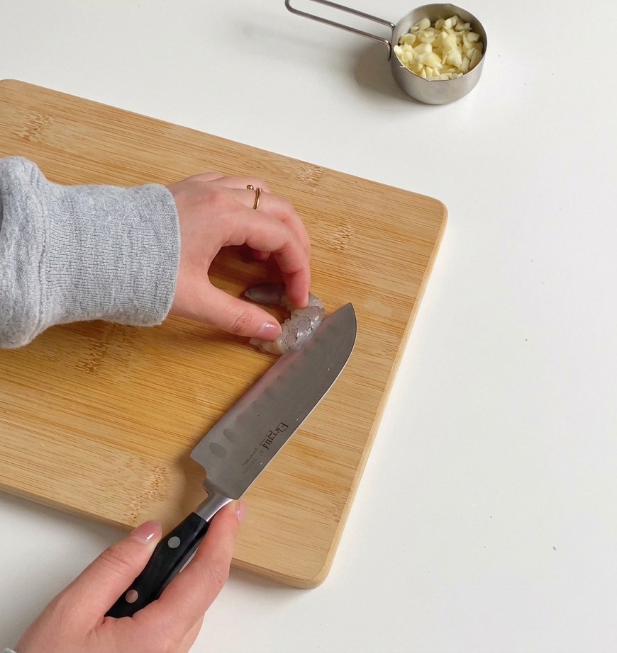 Close-up of hands using a sharp knife to butterfly a raw, peeled shrimp on a wooden surface.